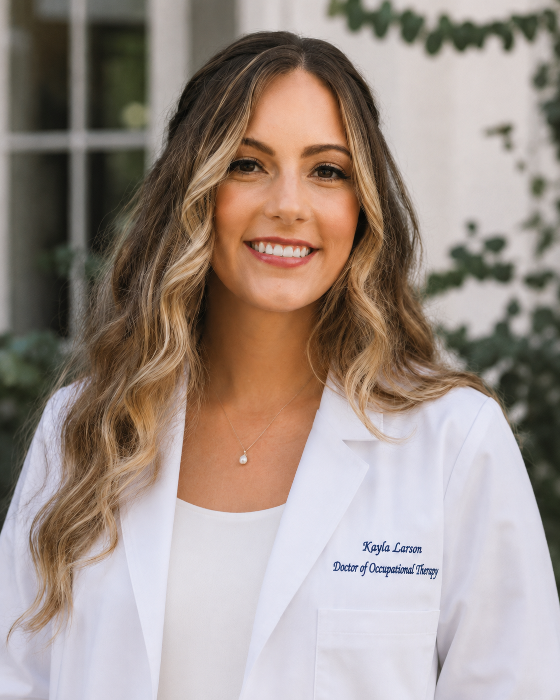 A woman smiling in a white coat with embroidered text that reads 'Kayla Larson, Doctor of Occupational Therapy', standing outdoors near greenery.