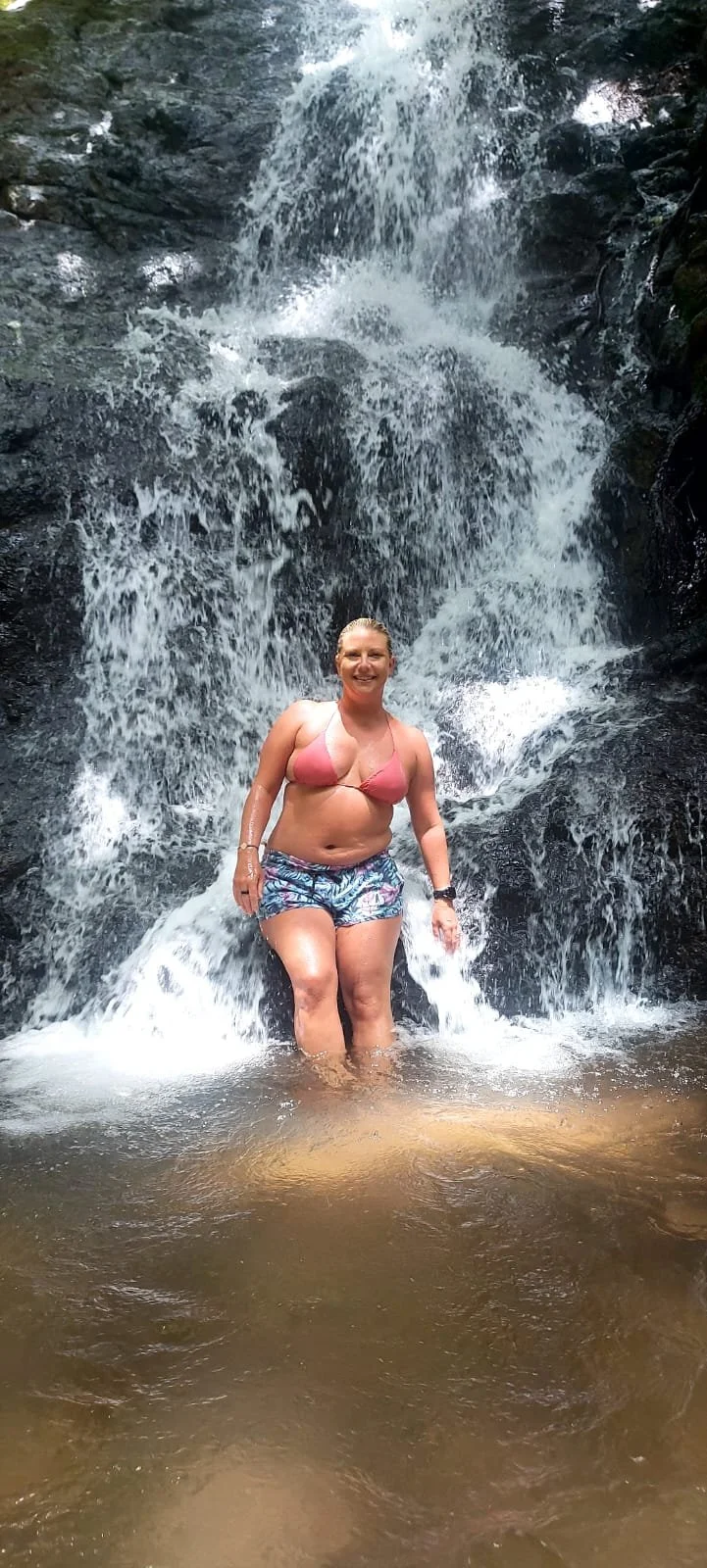Smiling woman in a bikini top and shorts standing in shallow water at a waterfall.