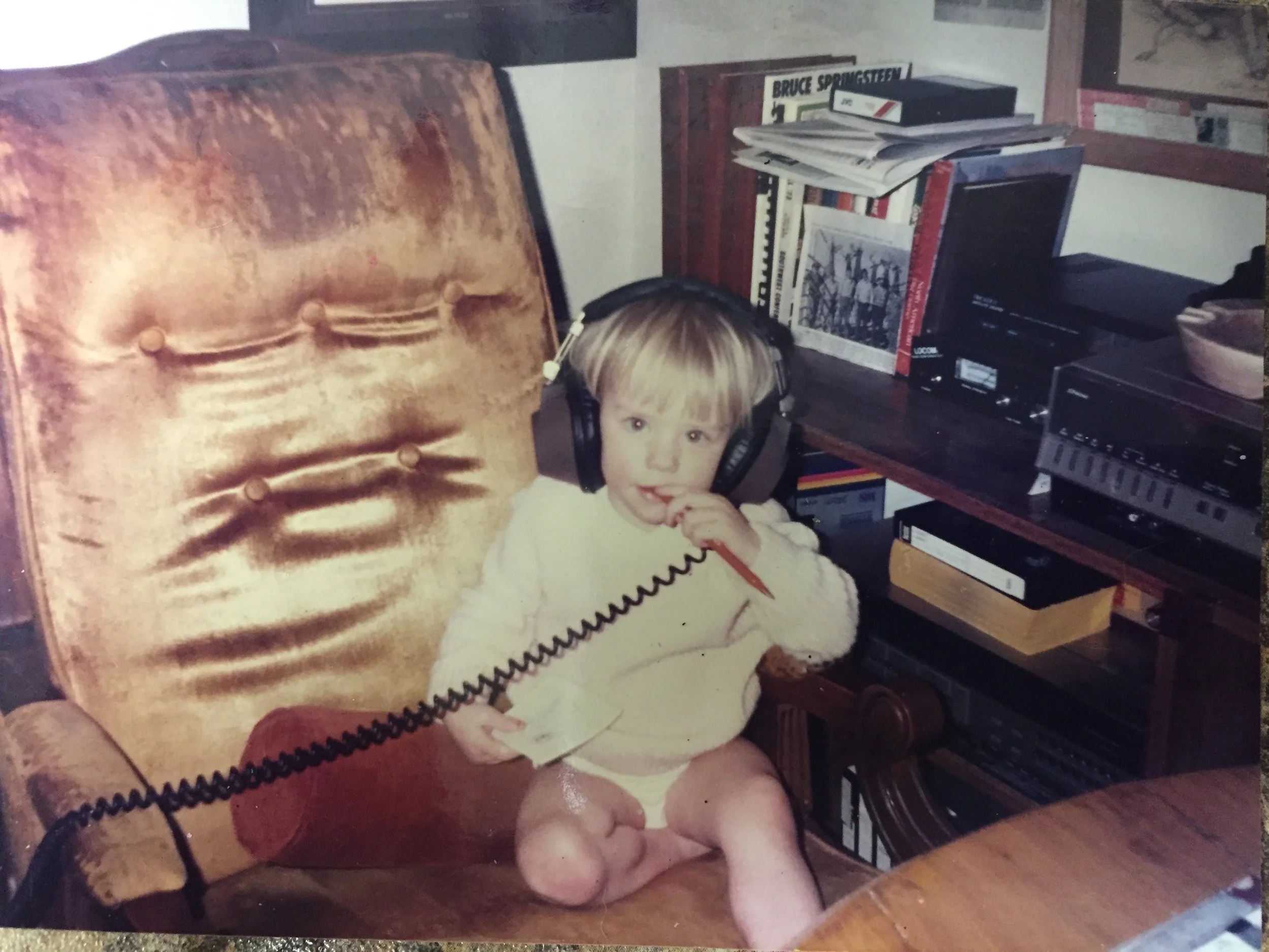 A young girl with light blonde hair and wearing headphones is sitting in a brown armchair while looking at the camera. There are shelves with books, magazines, and audio equipment behind her.