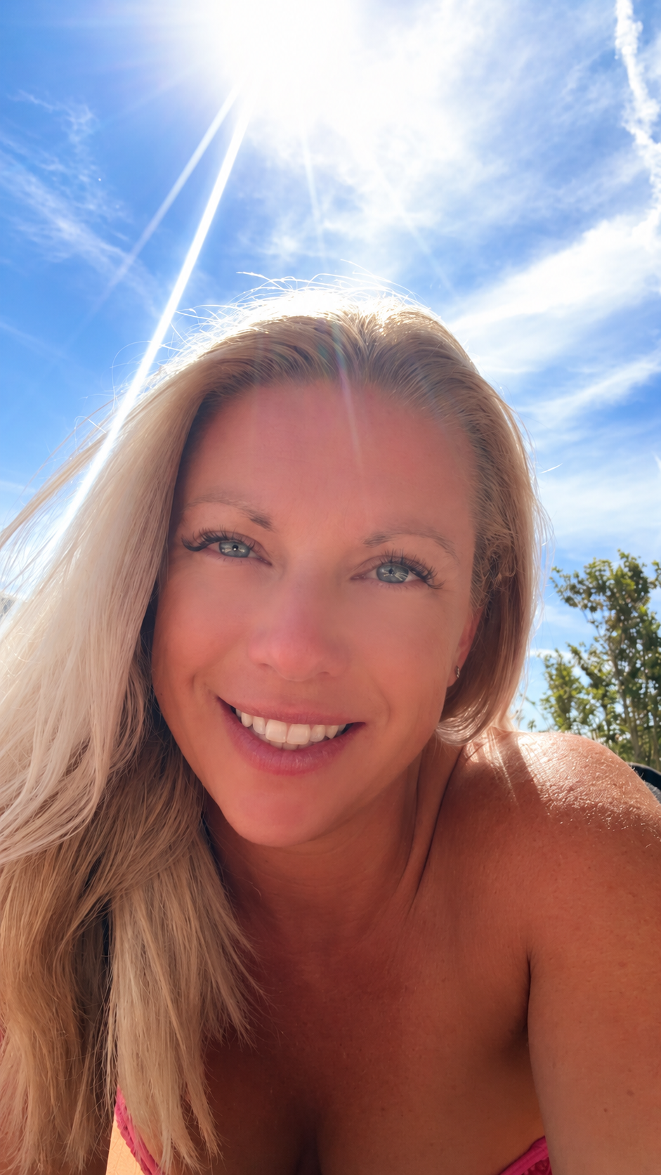 A smiling woman with long blonde hair outdoors under a bright blue sky with the sun shining brightly.