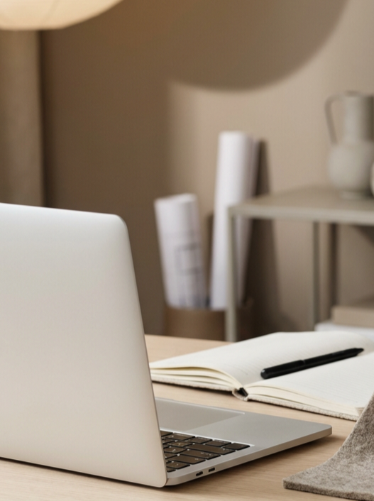 Open laptop computer, notebook, and pen on a desk with a beige wall and a bookshelf with rolled papers in the background.
