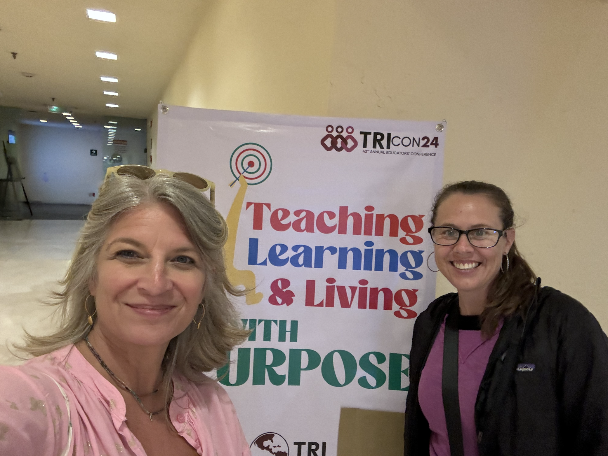 Two women taking a selfie at a conference with a banner that highlights 'Teaching, Learning, & Living' and the TRI CON 24 logo in the background.