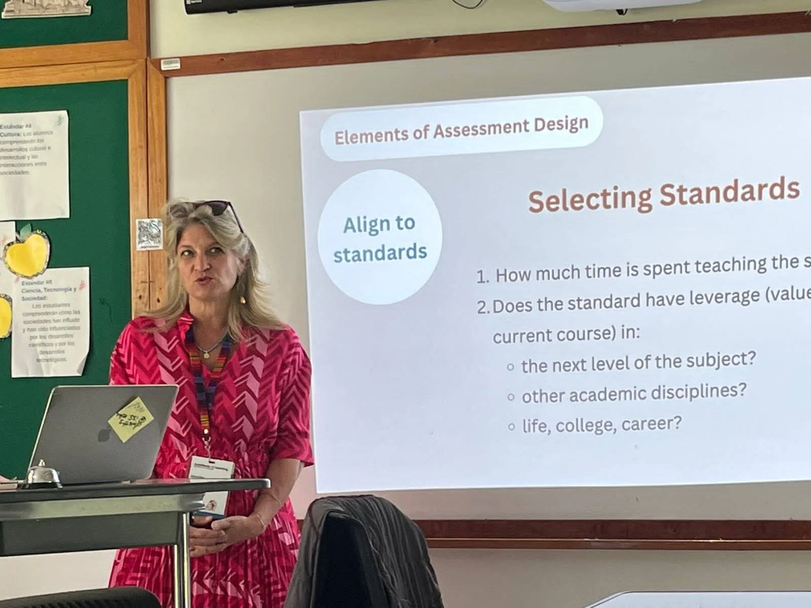 A woman with blonde hair wearing a pink patterned dress and sunglasses on her head stands in front of a presentation slide titled 'Elements of Assessment Design.' She is near a laptop and appears to be speaking or presenting, with classroom surroundings including a green bulletin board and posters in the background.