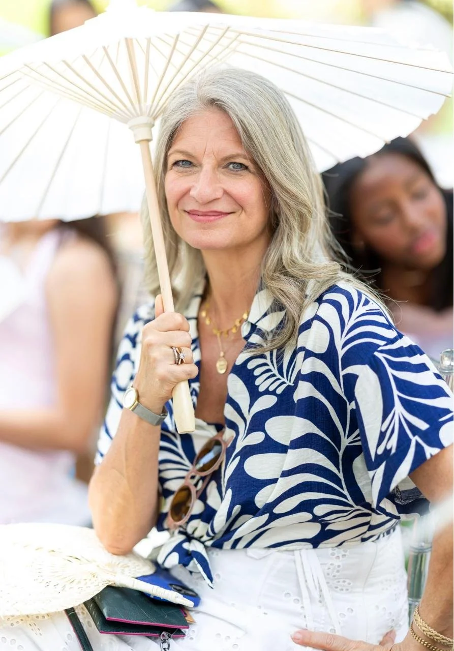 A woman with long gray hair, wearing a blue and white patterned blouse, holding a white parasol, smiling outdoors. She has sunglasses hanging from her blouse, a gold necklace, and jewelry on her wrist and finger. Two women are blurry in the background.