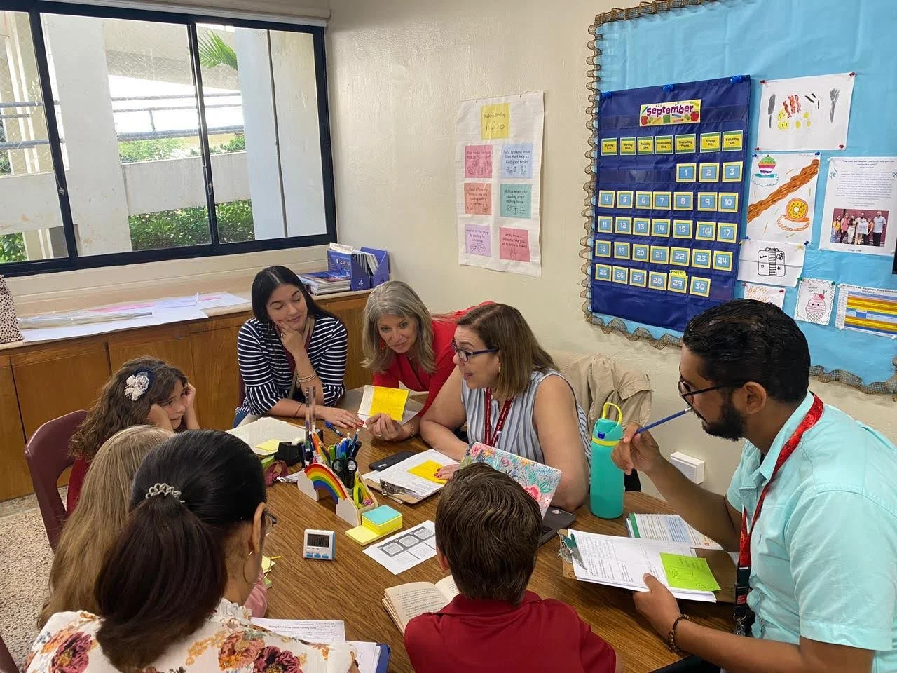 Parents and teacher engaged in a parent-teacher meeting in a classroom, sitting around a table with educational materials and papers, with colorful posters and a calendar on the wall.