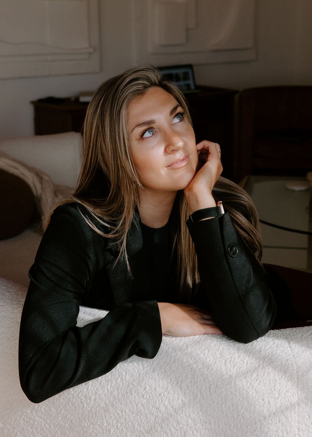 A young woman with long brown hair and blue eyes resting her chin on her hand while lying on a bed in a cozy room.