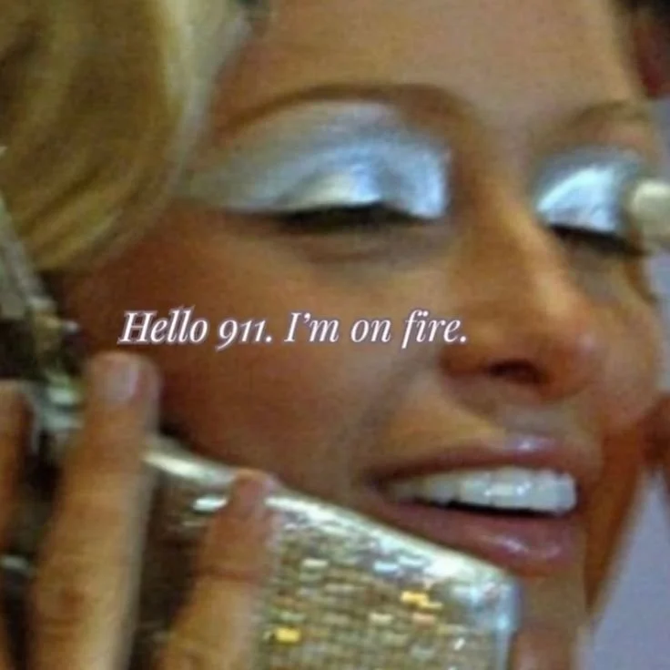 Close-up of a smiling woman with silver eyeshadow talking on a phone. Text overlay reads: 'Hello 911. I'm on fire.'