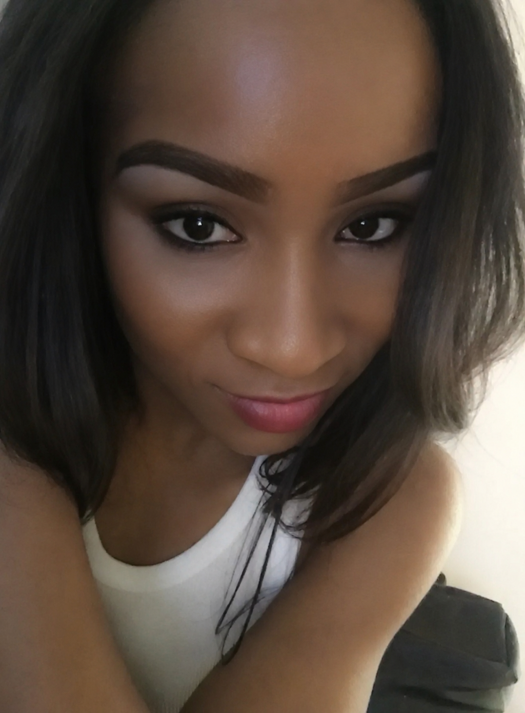 Close-up selfie of a young woman with dark hair, prominent eyebrows, and makeup, wearing a white sleeveless top.