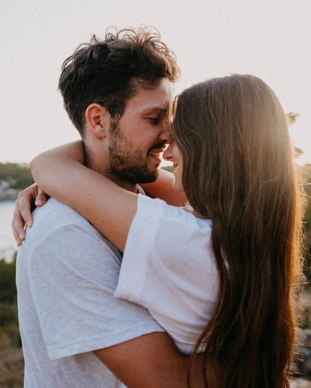 A couple embraces outdoors during sunset, touching foreheads and smiling at each other.