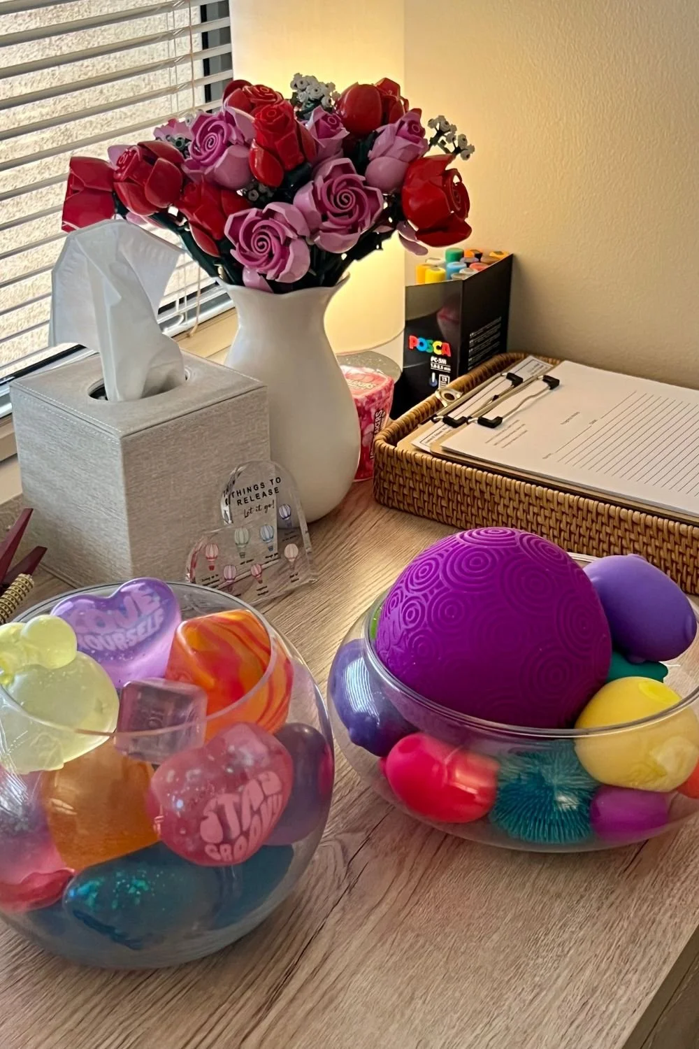 A desk holds a large white vase with pink and red roses, two bowls filled with colorful toys.