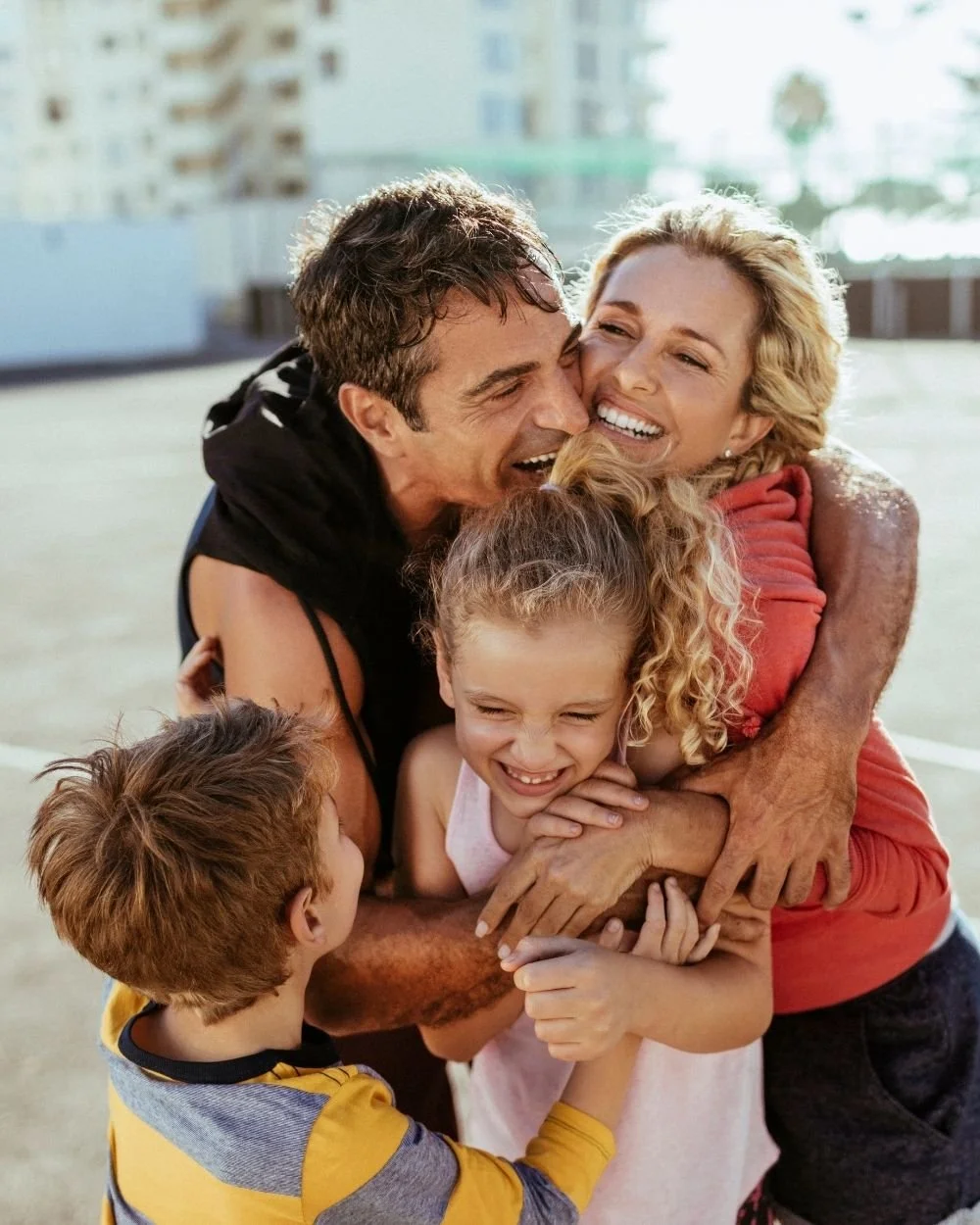 A happy family of four, including a man, woman, and two children, share a group hug outside on a sunny day, smiling and laughing.