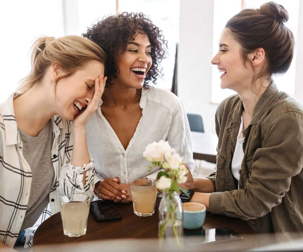 Three women sitting together at a table, sharing a joyful moment with smiles and laughter.