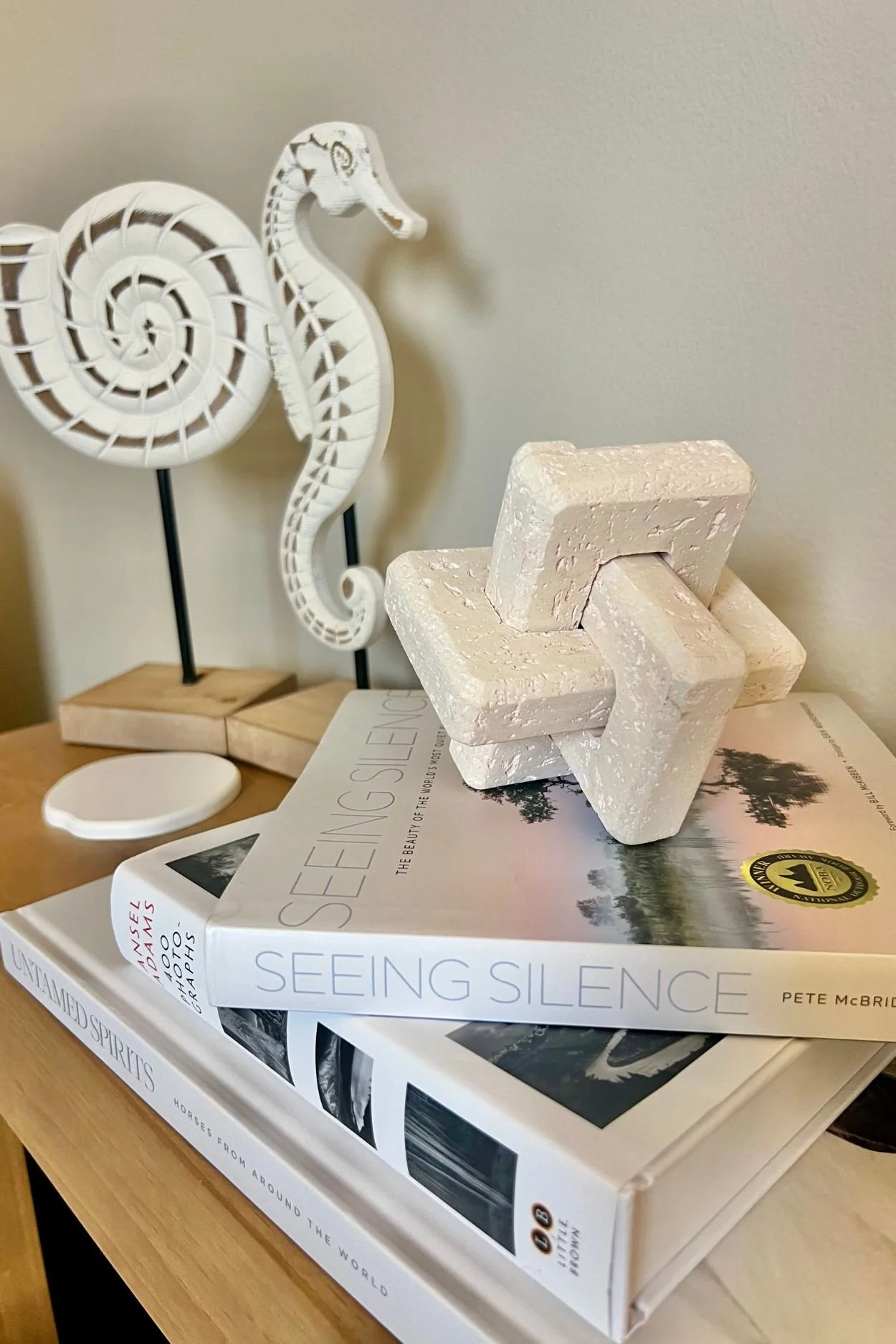 A close-up of a decorative white seahorse sculpture and a white abstract geometric sculpture placed on a table with a large book.