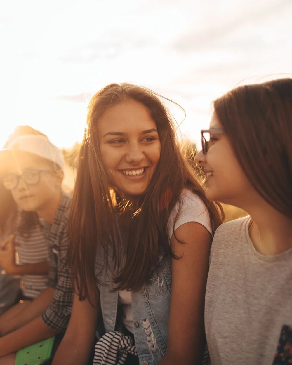 Three young women sitting outdoors during sunset, smiling and enjoying each other's company.
