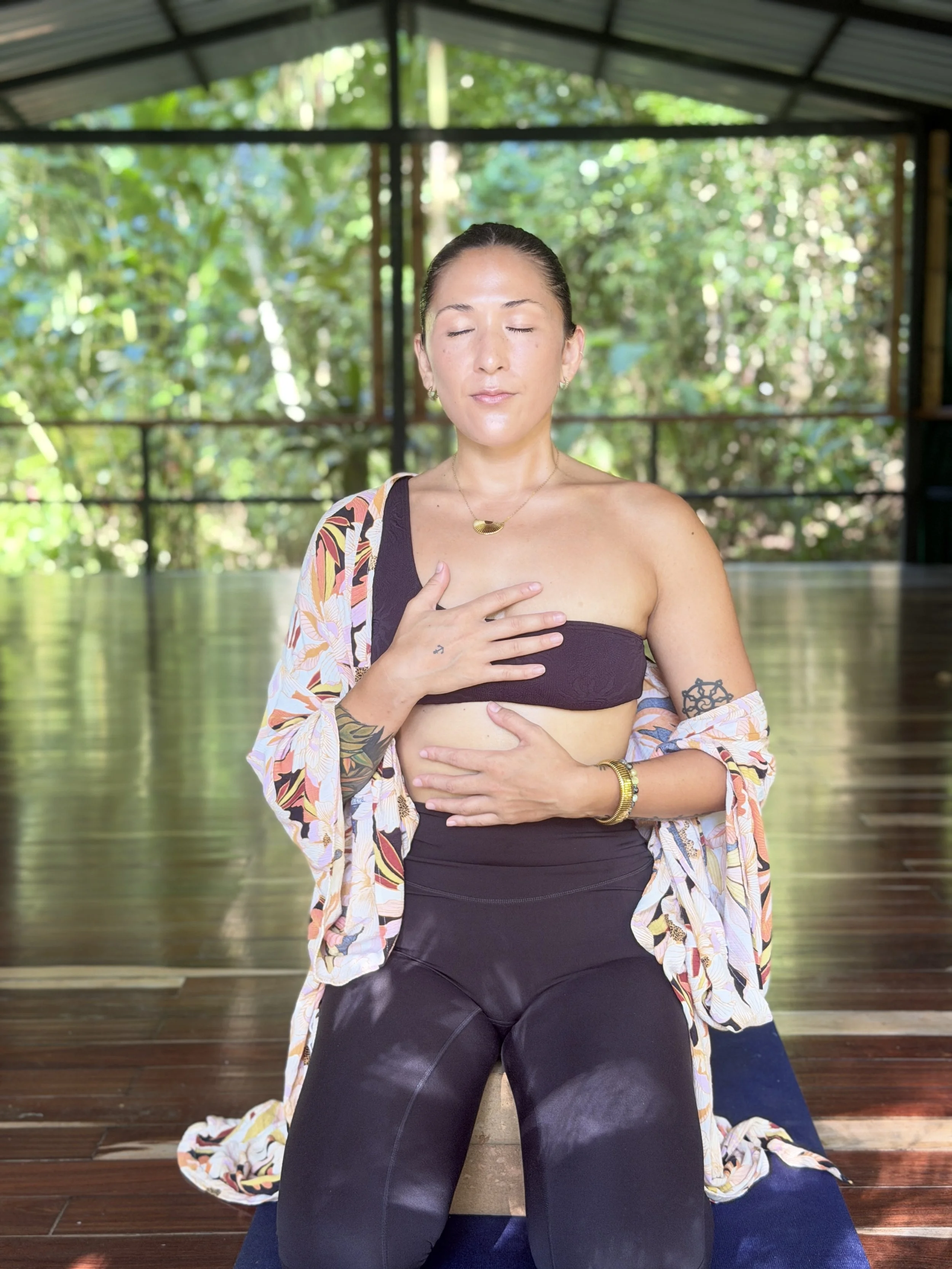 A woman with her eyes closed, sitting outdoors under a metal roof structure with lush green trees in the background. She is wearing a black strapless top, a colorful kimono-style jacket, gold jewelry, and has tattoos on her left arm. Her left hand is placed on her chest, and her right hand is on her stomach, in a meditative pose.