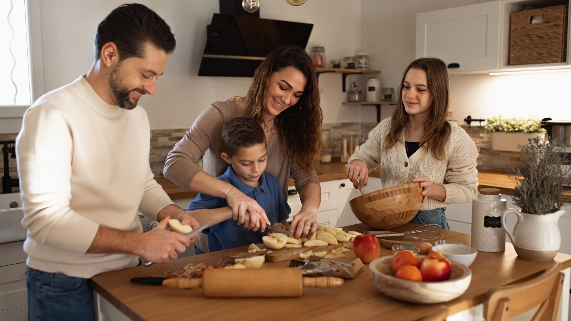 A family of four in a kitchen preparing and slicing fruit together. The father is on the left slicing an apple, the mother is in the center helping with the fruit, a young boy is between them, and a teenage girl stands on the right holding a wooden bowl, all smiling and engaging in a shared activity.