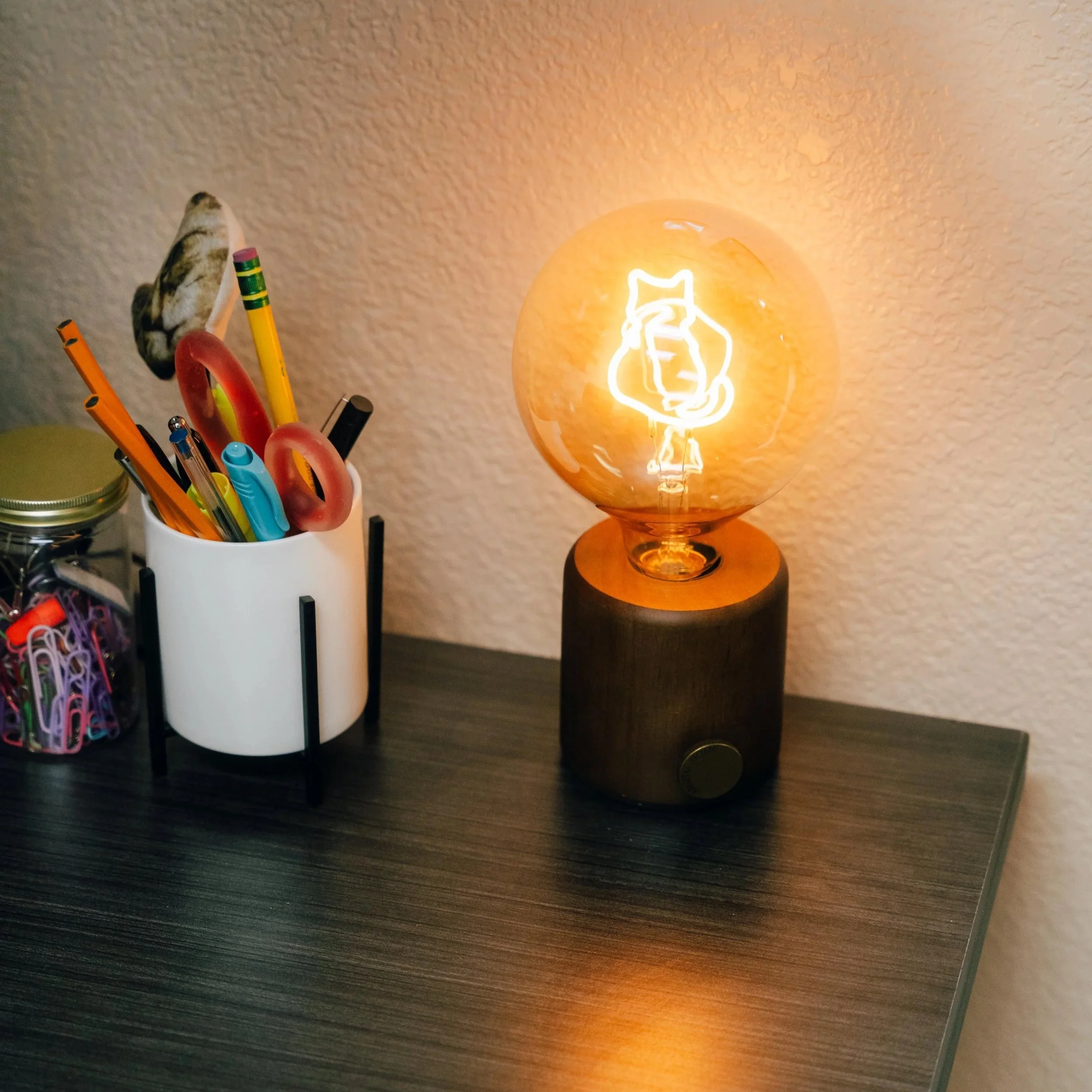 A decorative lamp with a neon outline of a bear holding a fish on a wooden surface next to a white container holding various colored pens and paper clips.