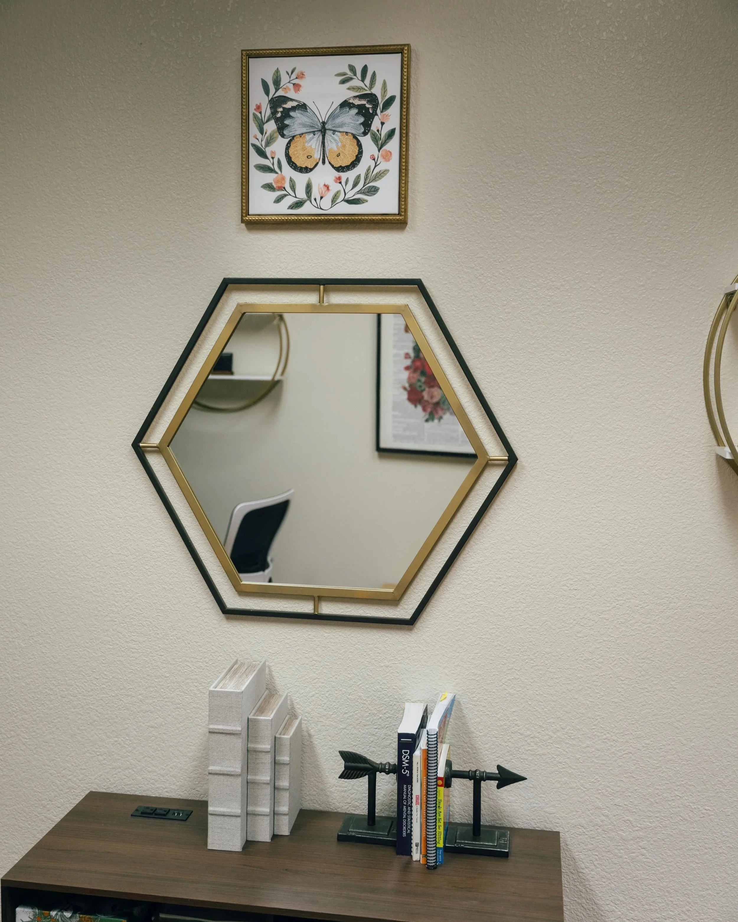 Decorative items on a wooden table, including white books, a black book, a black table lamp, and a black metal book stand, with a mirror, framed artwork of a butterfly, and a framed print on a beige wall in the background.