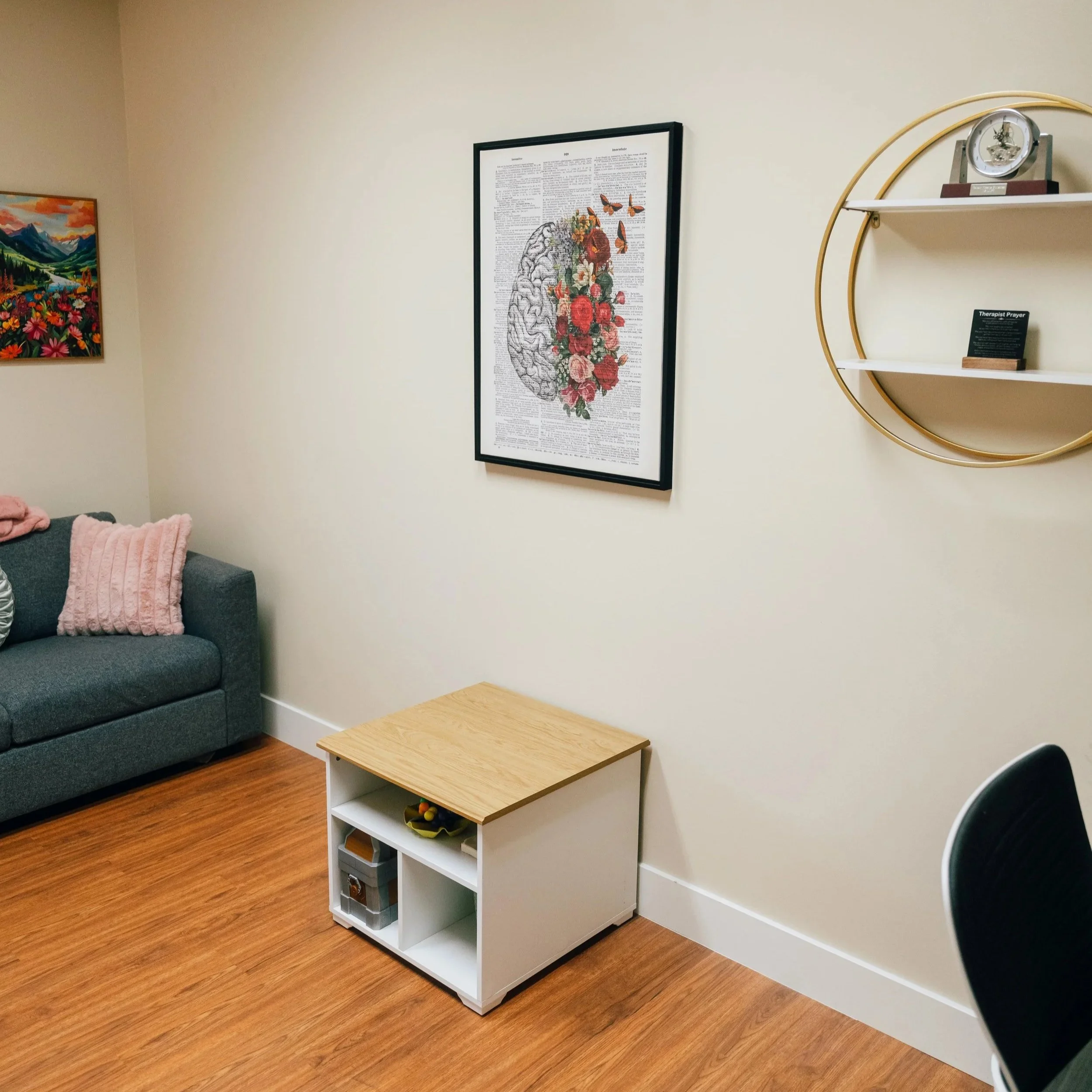 A small white bookshelf with four compartments, holding various items, is placed against a beige wall in a room with wood flooring. On top of the bookshelf, there is a small green plant and a black electronic device. The wall behind has two framed pi