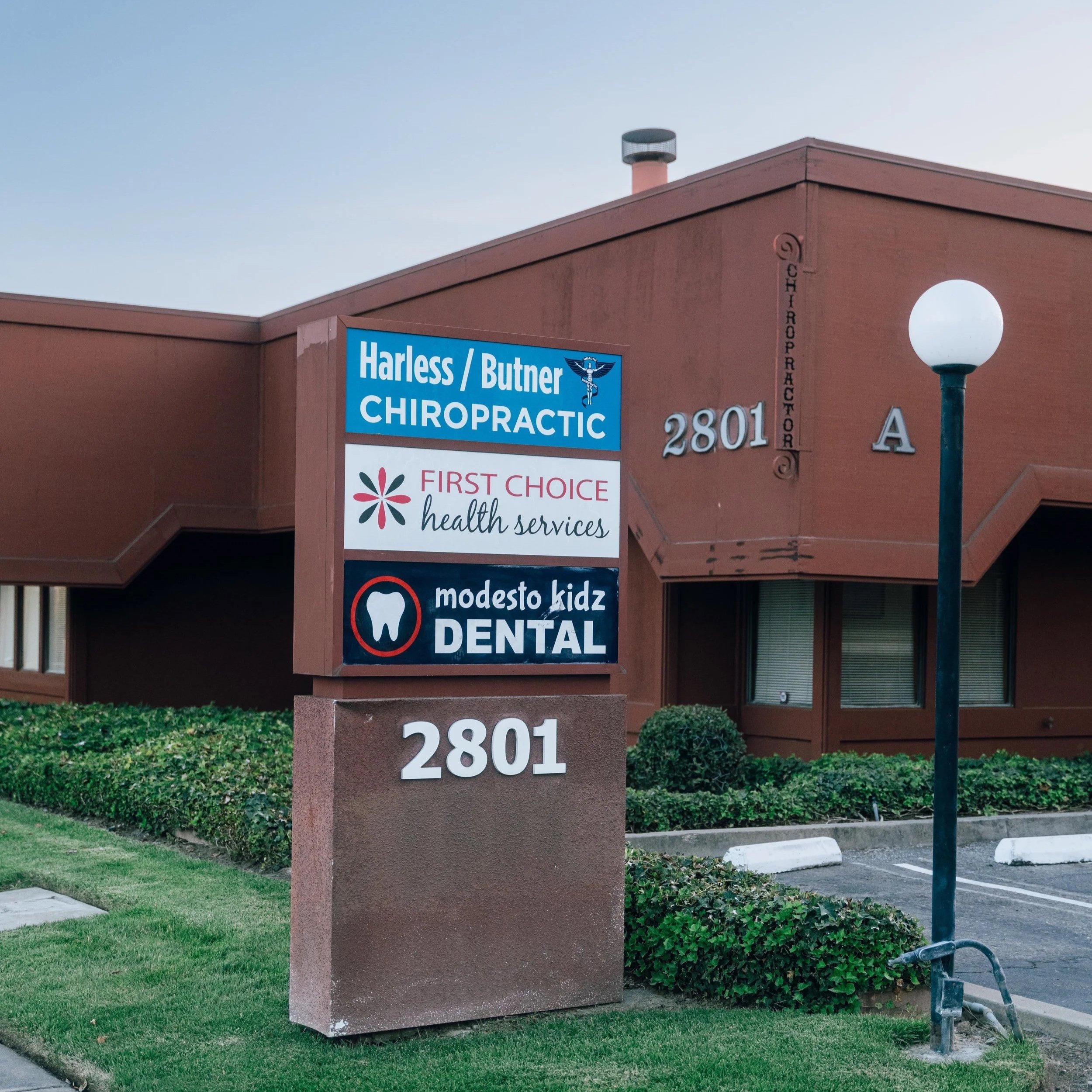 A building corner displays a blue sign advertising Harless / Butner Chiropractic, offering health services, with other health-related signs beneath. The building has a reddish-brown exterior, and there's a street lamp in front, along with a grassy ar