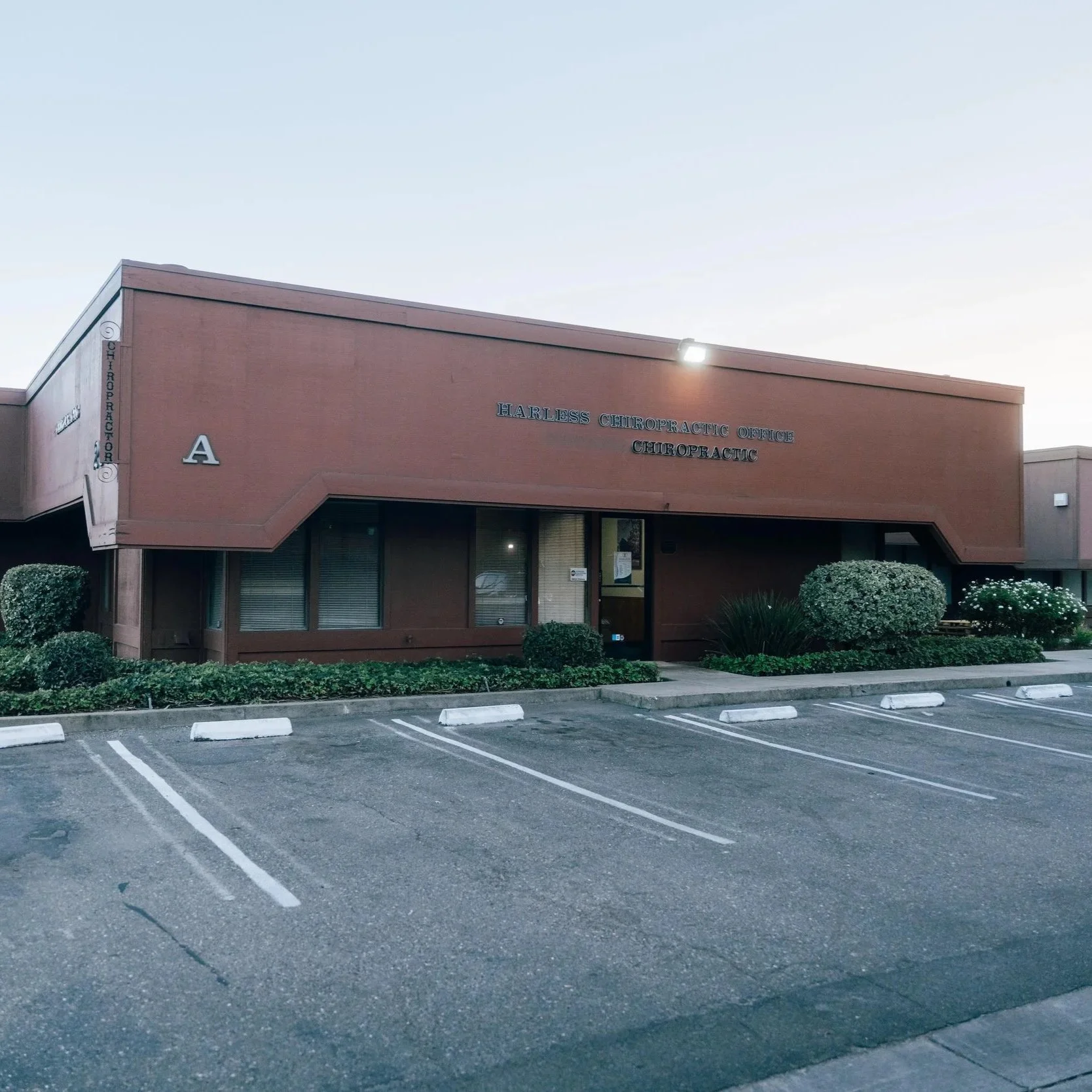 A single-story brick building with a pinkish hue, labeled as 'Harless Chiropractic Office' in black lettering. The building is numbered 'A' and has a parking lot in front with empty parking spaces and white striped lines, along with some greenery and bushes.