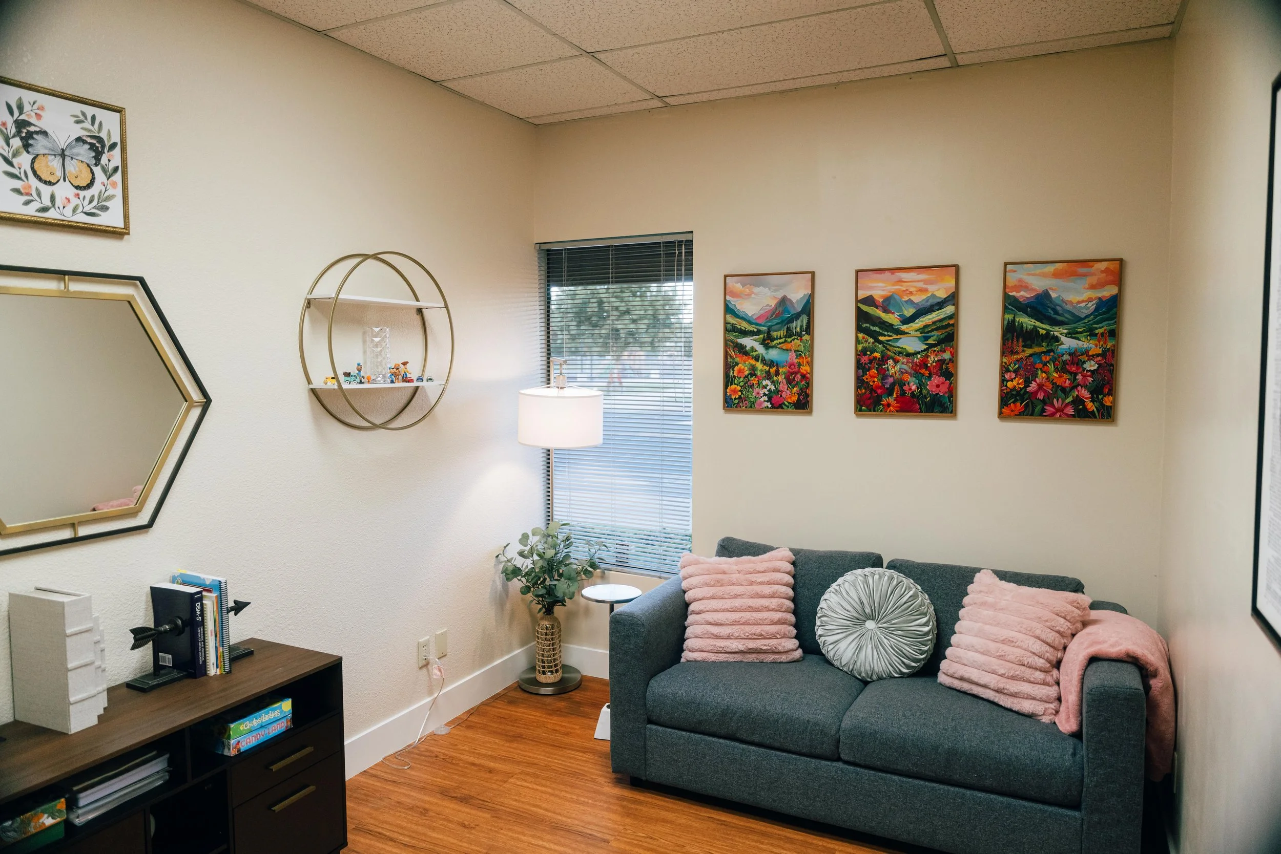 A cozy living room with a gray couch, pink and silver pillows, a wooden bookshelf, a white lamp, colorful landscape paintings on the wall, a window with blinds, and a small plant.