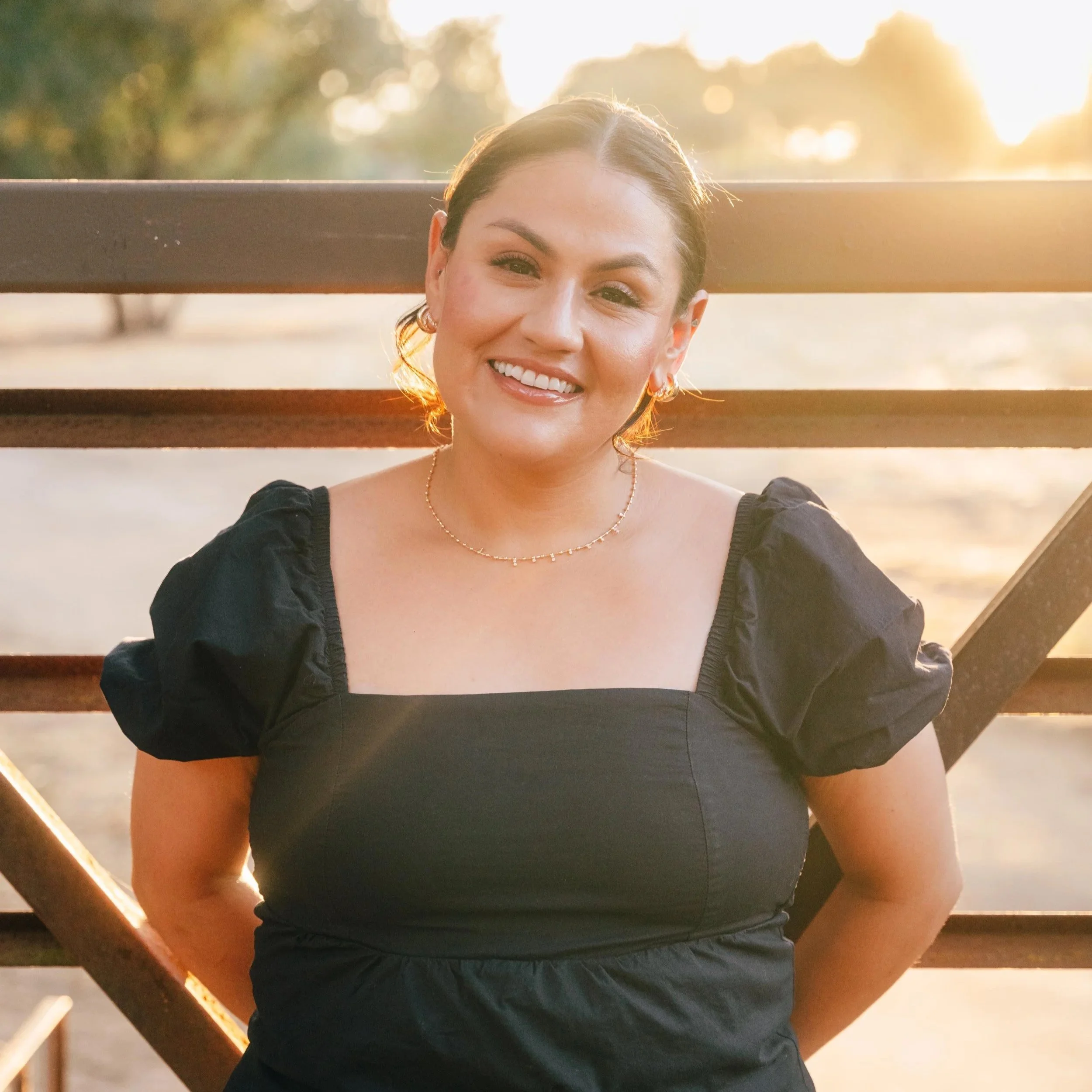 Smiling woman with gold earrings and a gold necklace sitting on a wooden bench outdoors during sunset, with trees in the background.