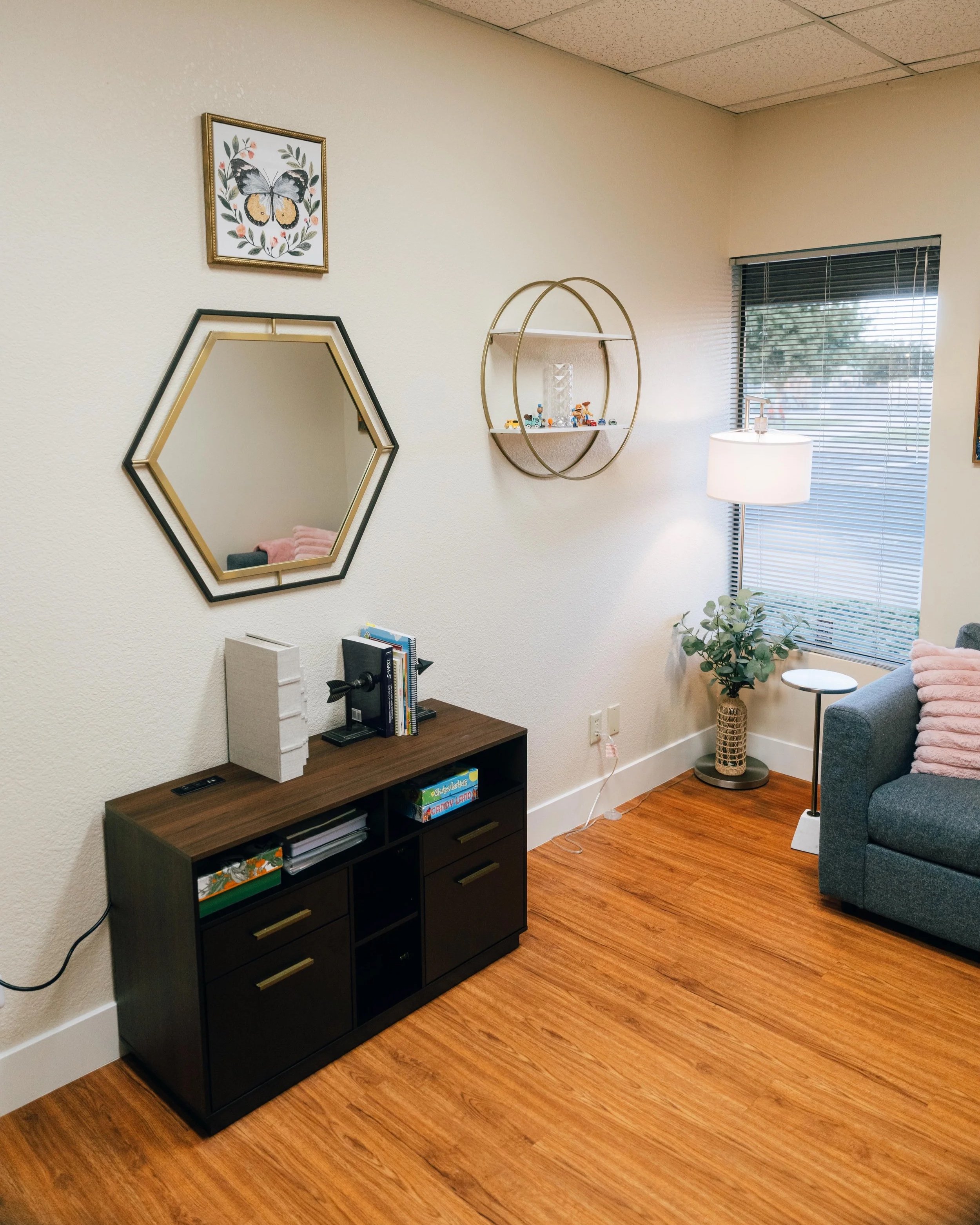 A corner of a room with a black bookshelf, a decorative mirror, a wall shelf with small toys, a potted plant, a floor lamp, a window with blinds, and a gray couch with pink pillows on a wood floor.