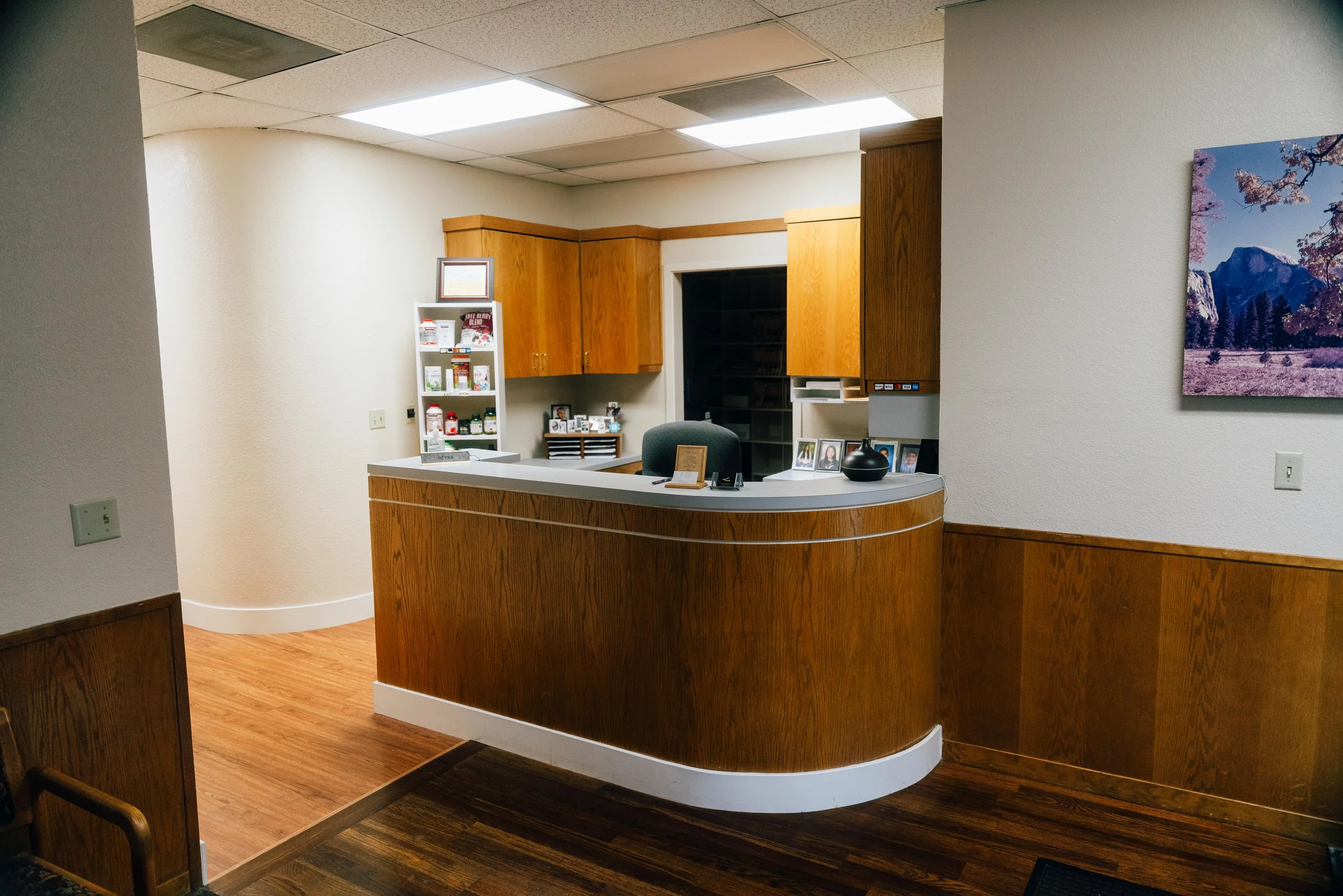 Interior view of an office reception area with a curved wooden reception desk, framed family photos on the desk, a white bookshelf with bottles and magazines, wooden cabinets, a wall with a landscape painting, and ceiling lights.