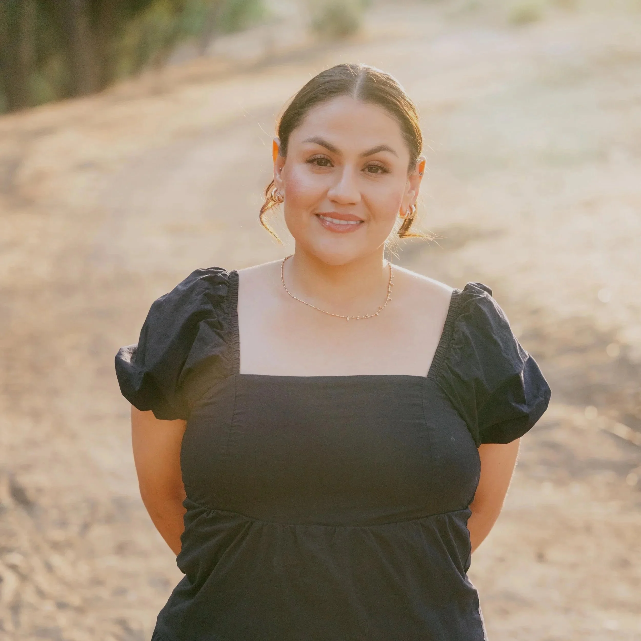 A young woman with dark hair pulled back, wearing hoop earrings and a black off-shoulder top, smiling outdoors with blurred background of sunlight and foliage.