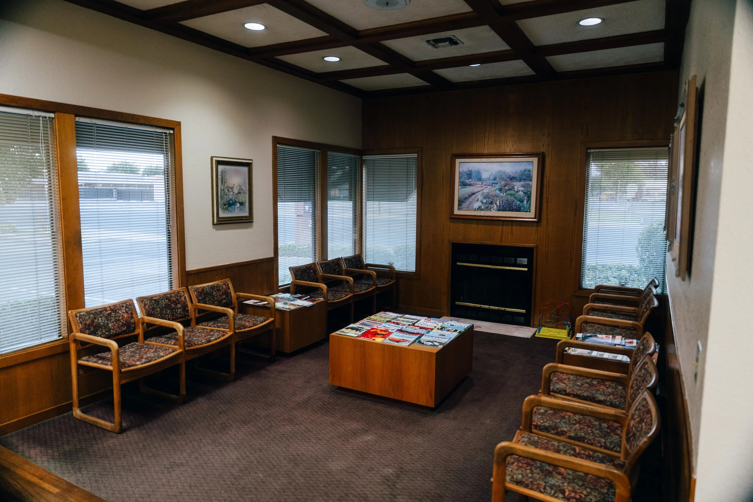 A waiting room with beige walls, wood paneled accents, and multiple chairs with floral upholstery arranged in rows. There are windows with blinds, framed landscape paintings on the walls, and magazines on a small table in the center. Recessed ceiling