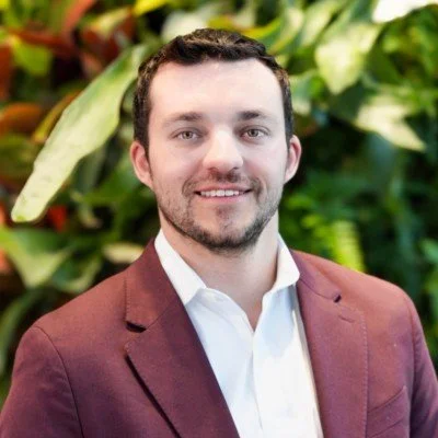 A man with short dark hair and a beard, wearing a white shirt and maroon blazer, smiling in front of a background of green plants.