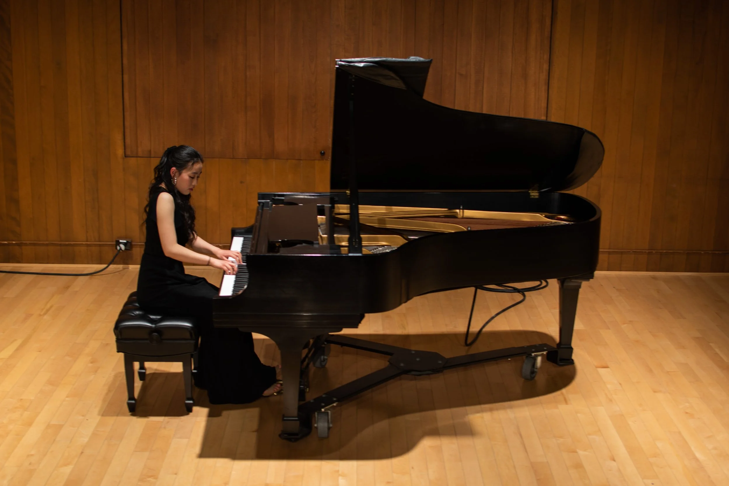 A young woman with long dark hair, wearing a black dress, plays a grand piano on a stage with wooden paneled walls.