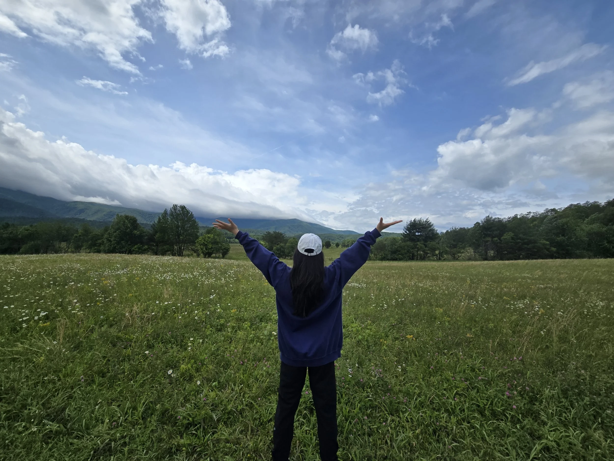 Child in a blue jacket and white cap standing in a grassy field with trees, mountains, and a partly cloudy sky in the background, arms raised in a welcoming gesture.