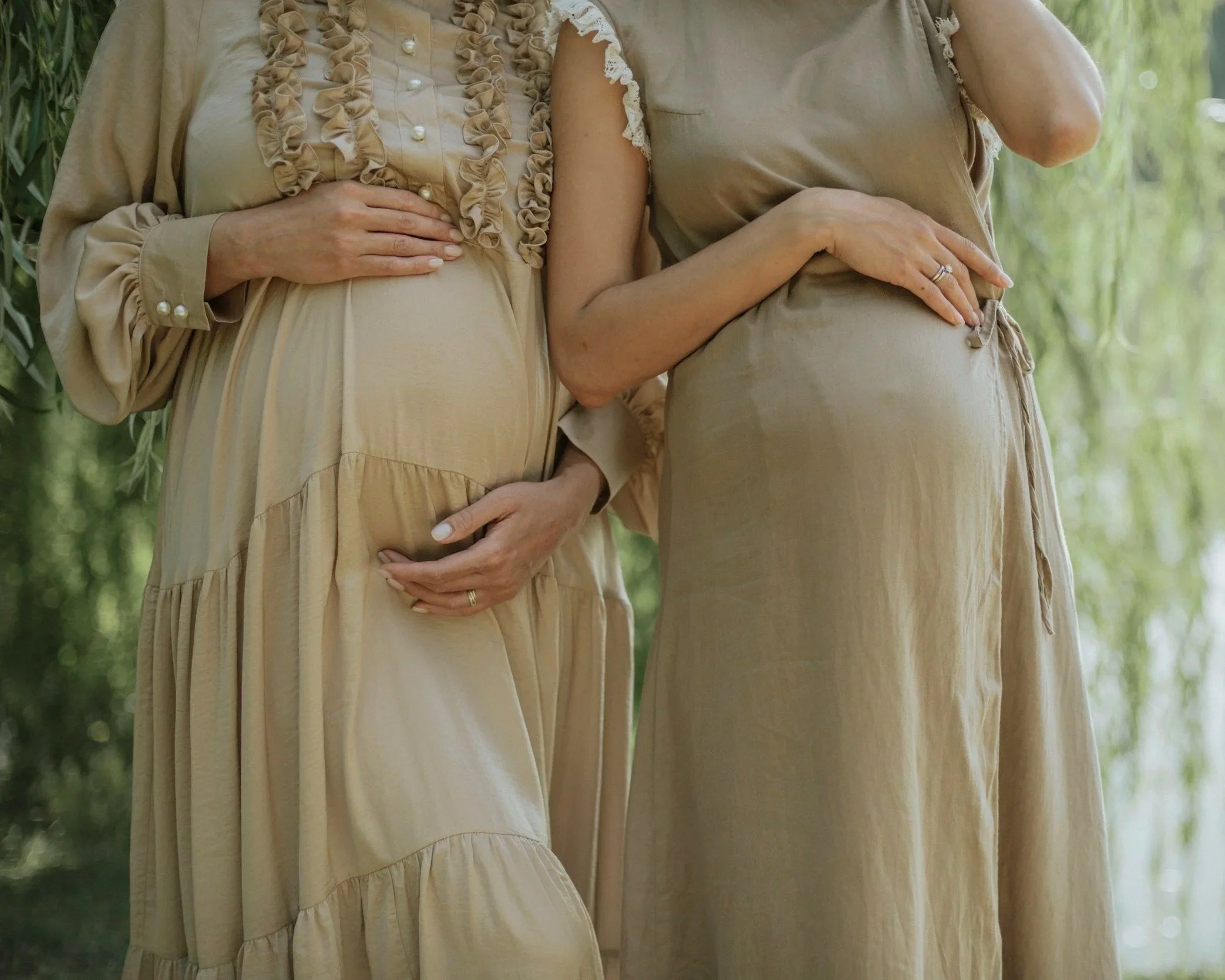 Two women standing close together outdoors, wearing beige dresses, with one woman resting her hand on her belly.