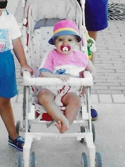A baby sitting in a stroller, wearing a pink outfit, a colorful hat, and a pacifier.