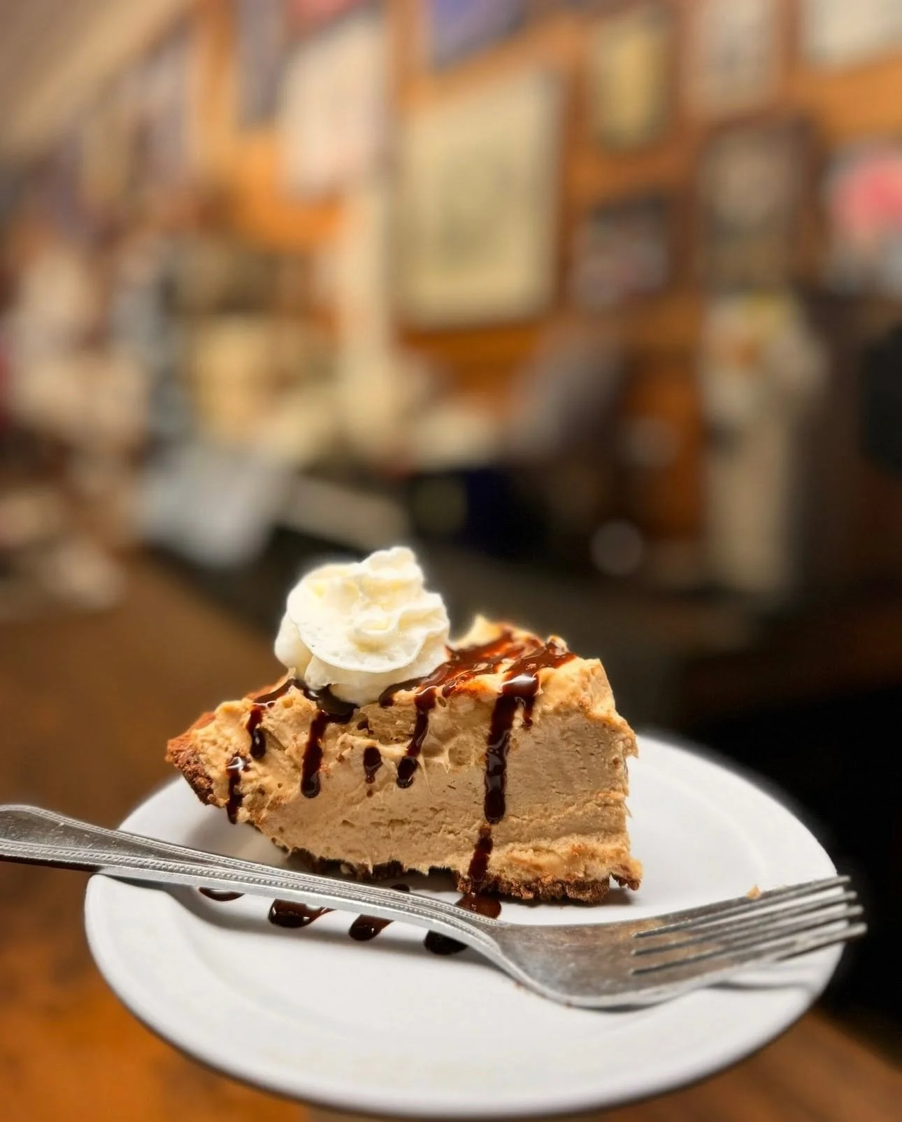 A slice of cheesecake topped with whipped cream and chocolate syrup on a white plate, with a fork resting on the plate, in a cozy bakery or cafe setting with wooden decor and framed pictures in the background.