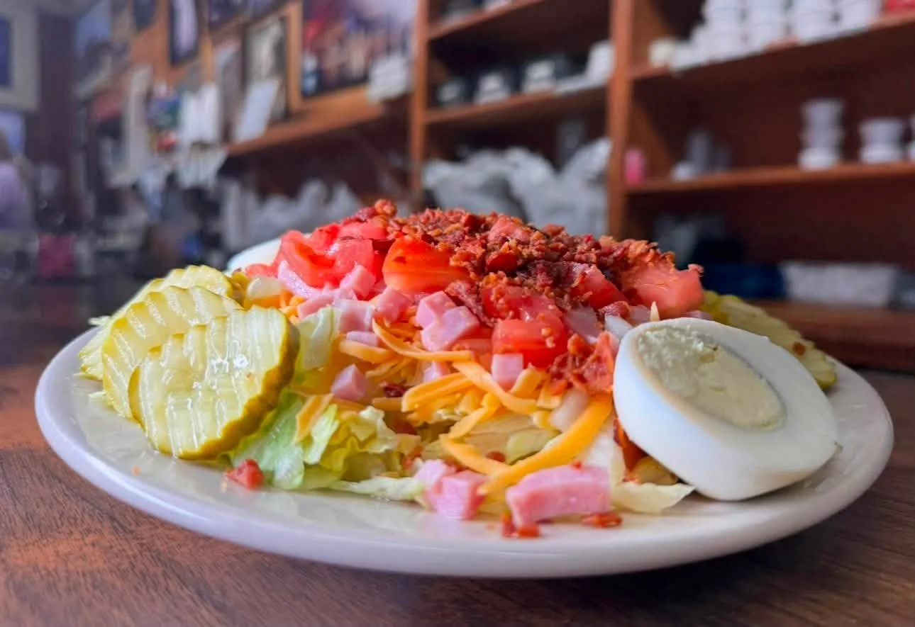A plate of comida corrida including sliced pickles, chopped lettuce, shredded cheese, diced ham, tomatoes, ground beef, and a sliced hard-boiled egg.