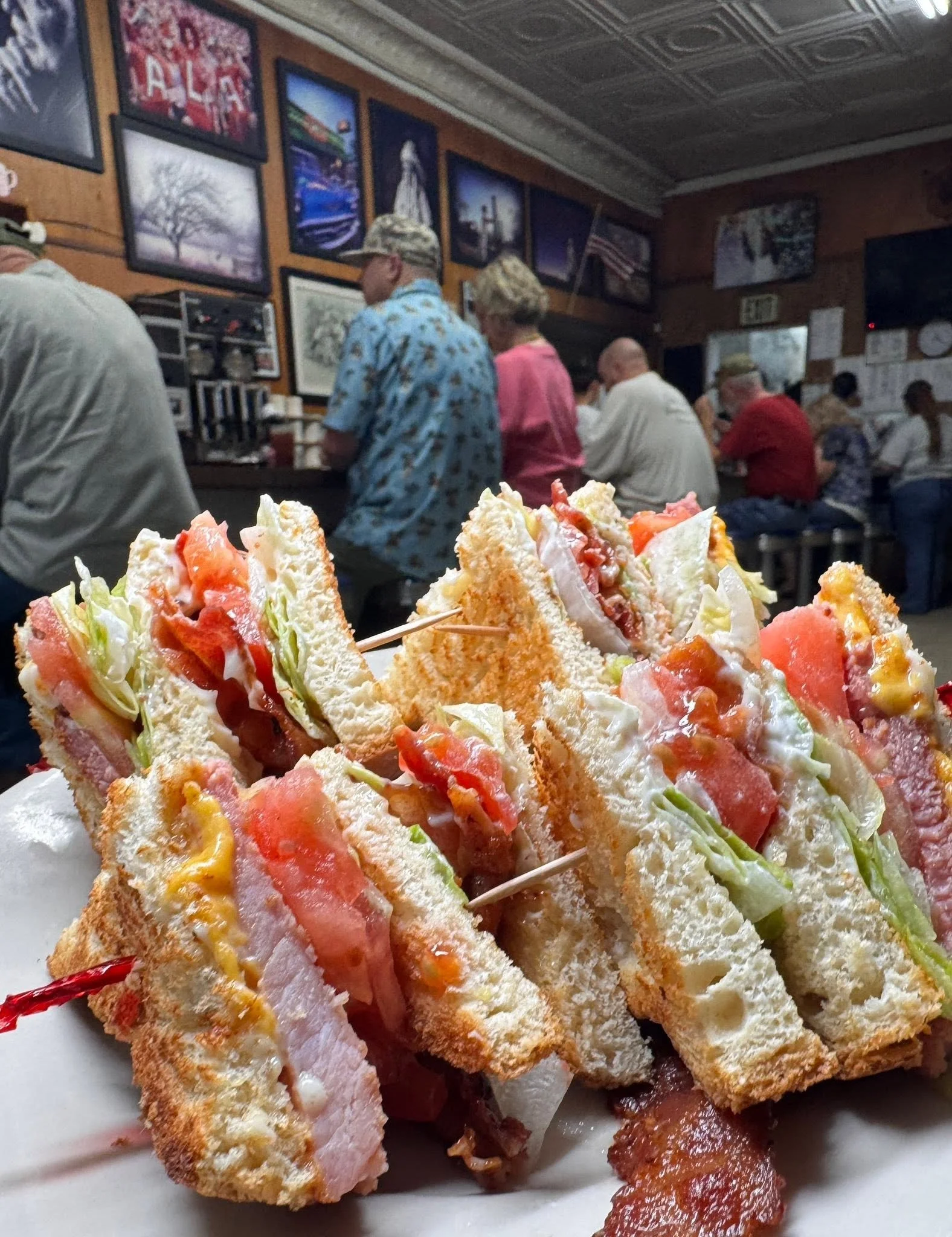 Close-up of next to a plate of layered sandwiches with various meats, cheese, lettuce, tomato, and condiments. In the background, there are customers sitting at tables inside a restaurant with artwork and pictures on the walls.