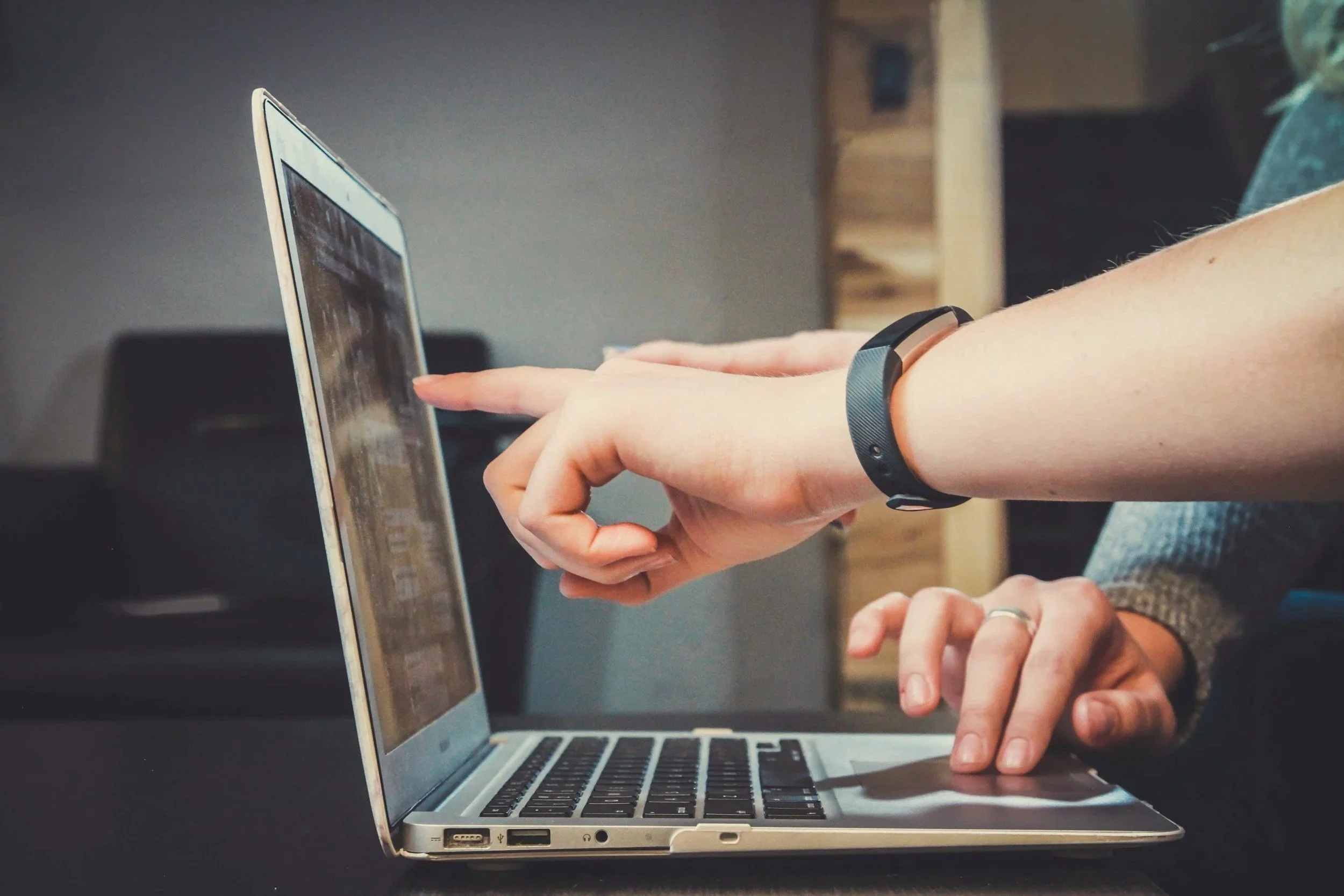 Close-up of a person's hand and arm as they use a laptop computer, with the person's right hand pointing at the screen and wearing a fitness tracker or smartwatch on their left wrist. The laptop is on a dark surface with a blurred background.