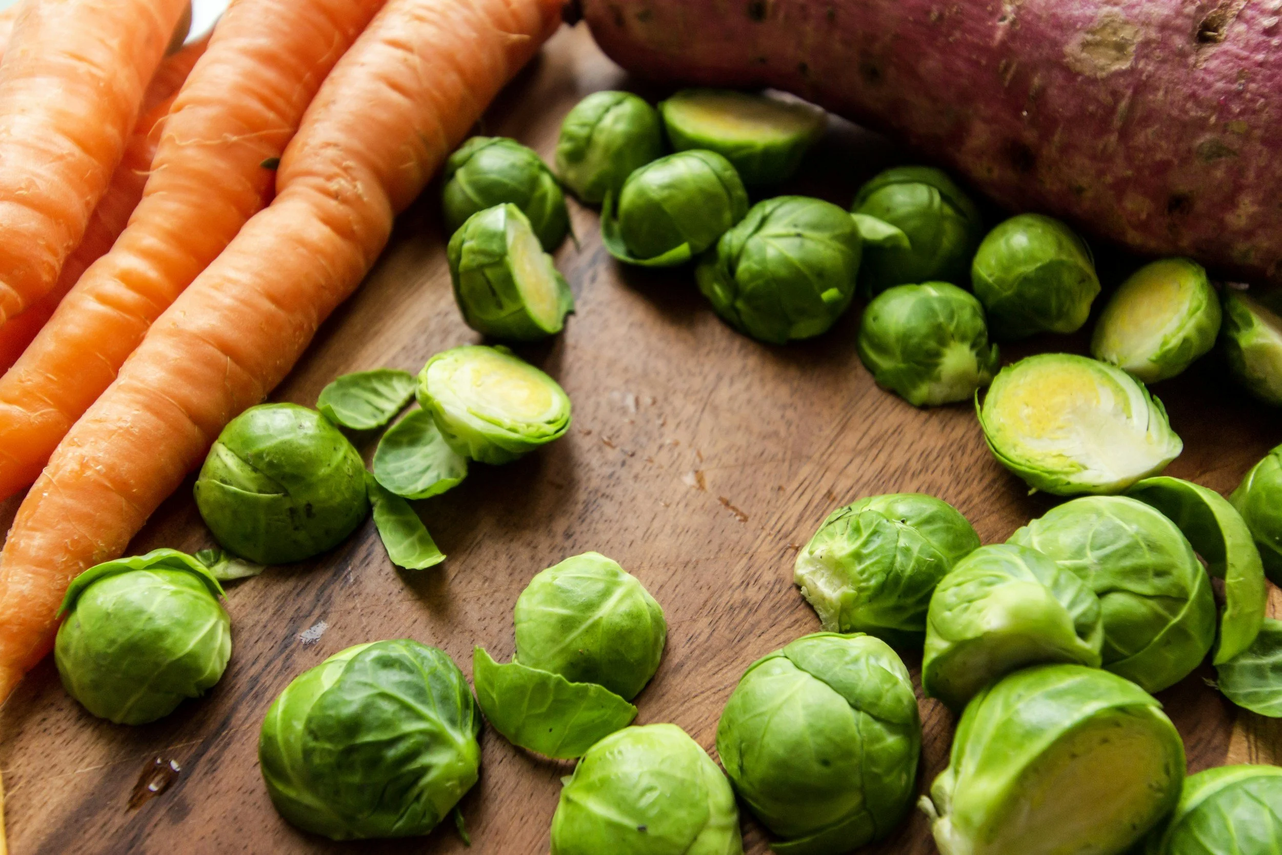 Carrots, Brussels sprouts, and a sweet potato on a wooden cutting board.