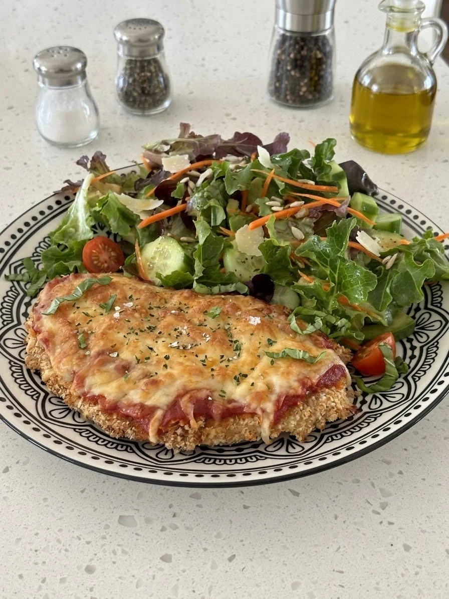 A plate containing a slice of breaded chicken Parma and a side salad with cherry tomatoes, cucumber, carrots, lettuce, and sunflower seeds, set on a decorative black and white plate. In the background, there are salt, pepper, and seasoning shakers, a bottle of olive oil, and a white countertop.