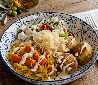 A bowl of Mediterranean food with vegetables, rice, falafel, and salad, garnished with sauce and herbs.