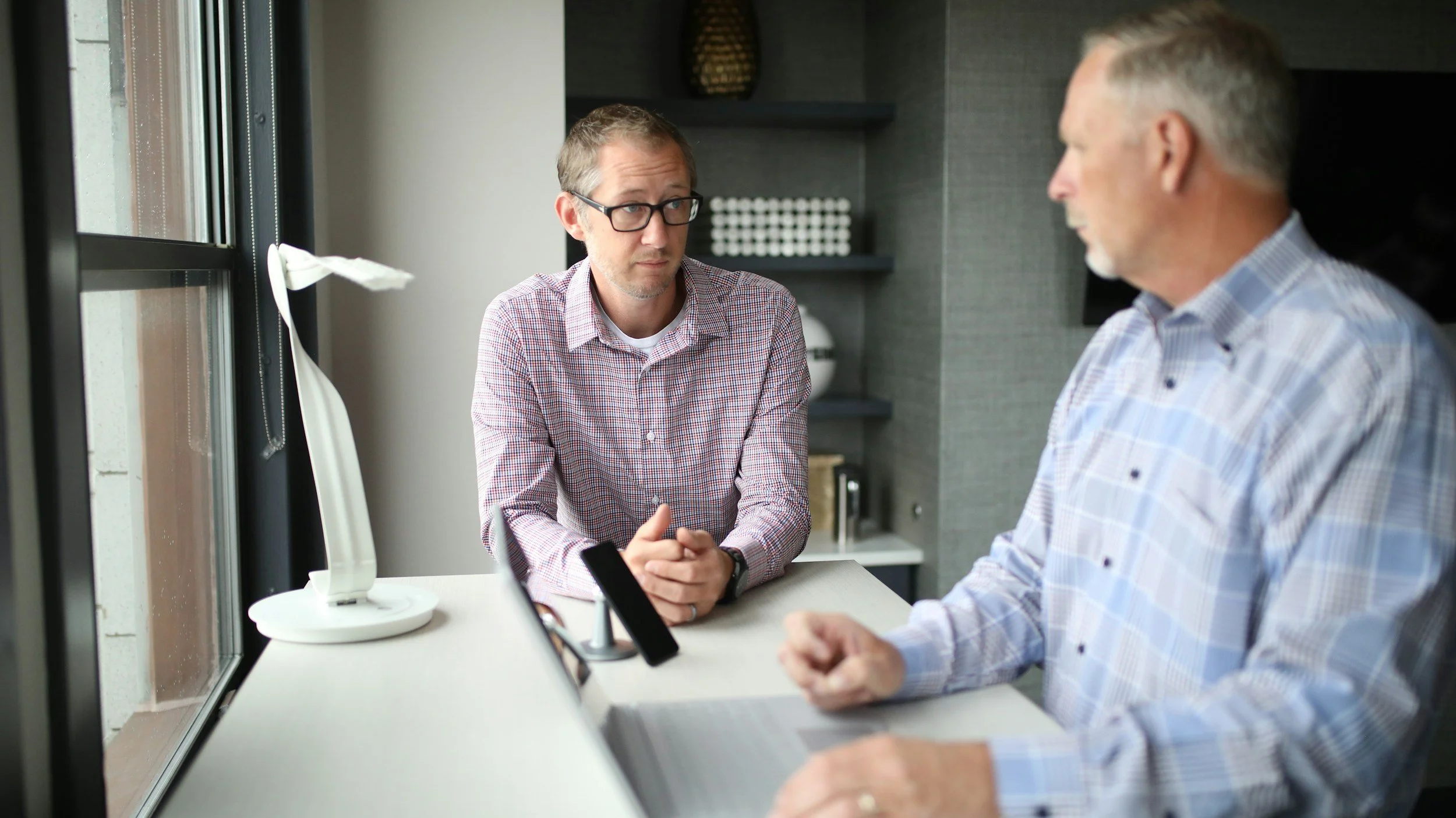 Two men having a conversation at a table in an office setting, with a window and some shelves in the background.