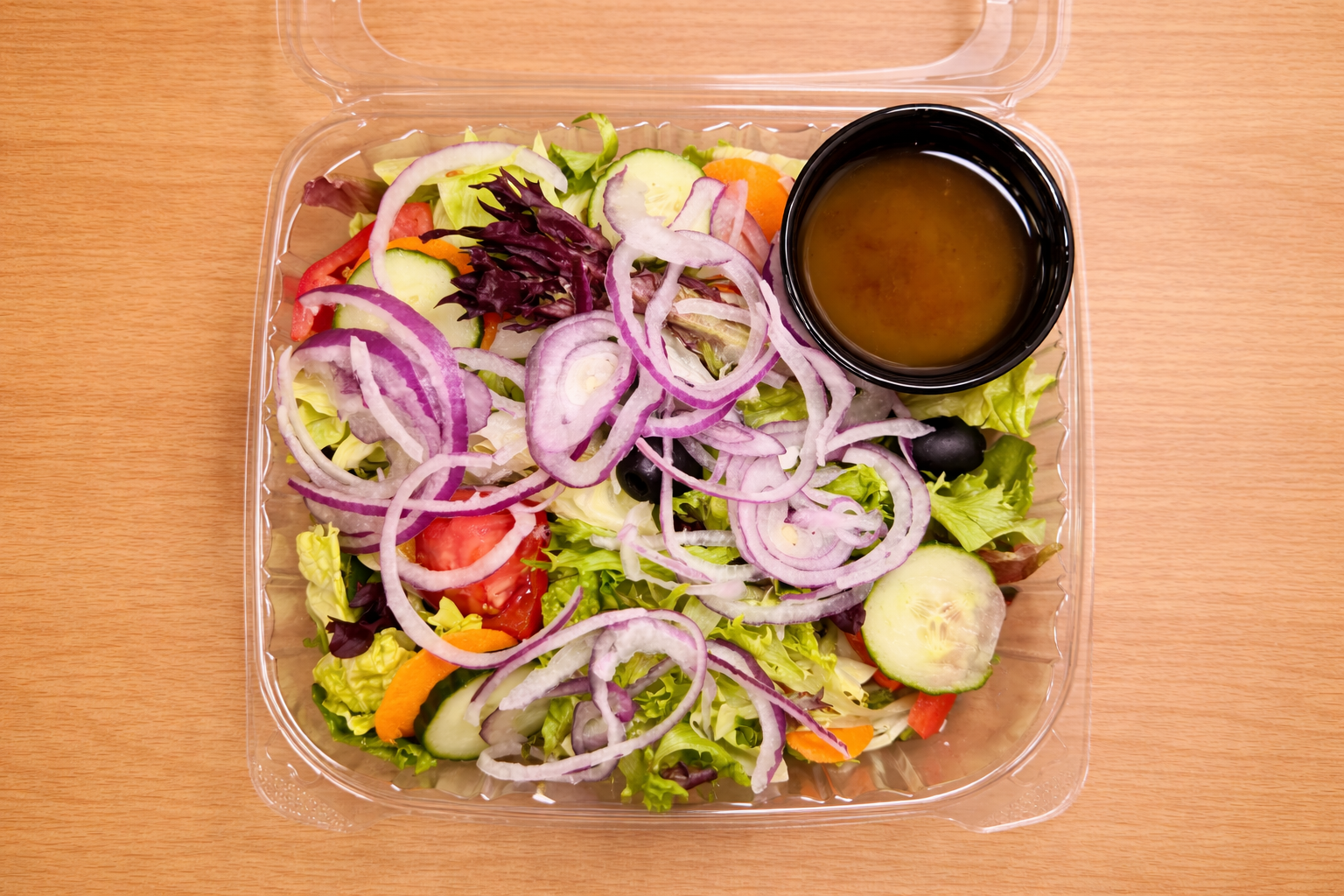 Fresh vegetable salad with lettuce, cucumbers, tomatoes, red onions, carrots, black olives, and a side of dressing in a small black cup
