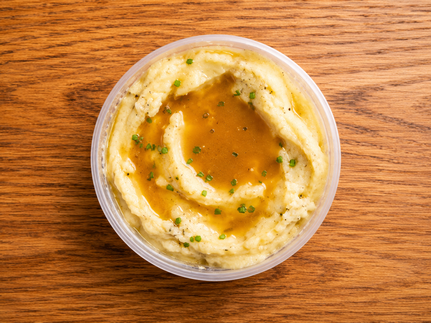 A bowl of mashed potatoes topped with gravy and sprinkled with chopped chives, on a wooden surface.