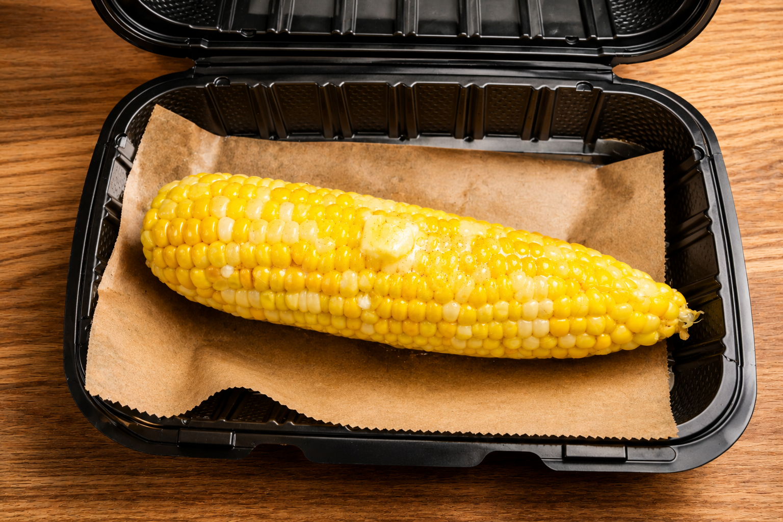 Fresh corn on the cob inside a black plastic container lined with brown paper, on a wooden surface.