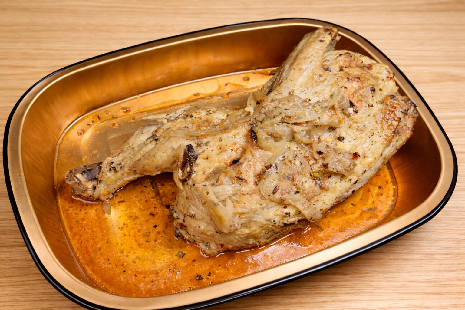 Baked chicken leg with onion bits in an orange sauce in a gold and black tray on a wooden surface.