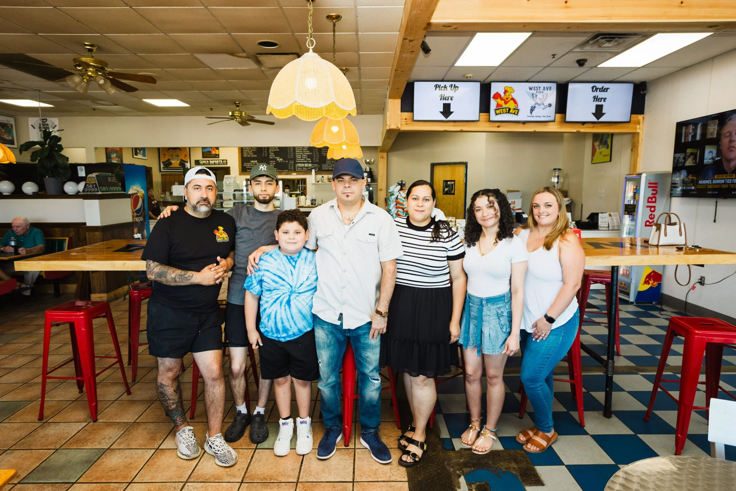 Group of seven people posing inside a restaurant near a red table with a light-colored tiled floor and a menu board behind them. Three digital screens, a water dispenser, and a vending machine are also visible.
