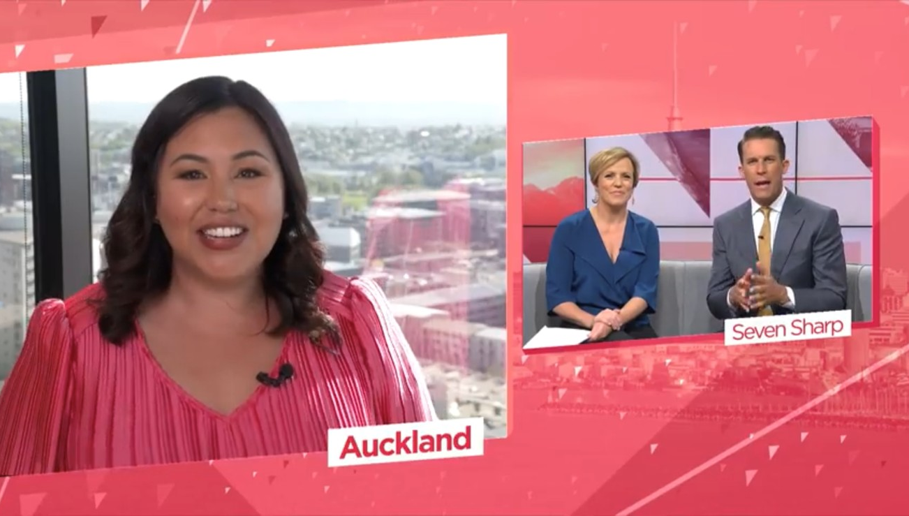 A woman smiling at the camera, wearing a pink striped top, with a cityscape view behind her. On screen, there are two smaller images: a woman in blue sitting next to a man in a suit, with the name "Seven Sharp" written below, and her own image with the caption "Auckland".